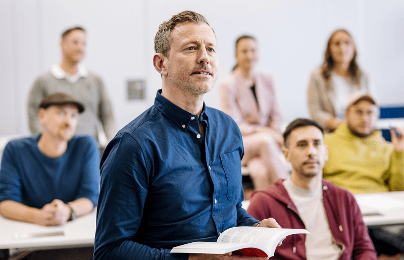 Teilnehmer eines VdS-Firmenseminars mit aufgeschlagenem Fachbuch spricht im Dialog mit Kolleginnen und Kollegen – praxisnahe, individuell zugeschnittene Weiterbildung im eigenen Teamumfeld.