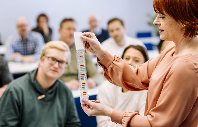 Dozentin erklärt im Seminar zum organisatorischen Brandschutz die Funktion farbiger Glasfässchen an Sprinkleranlagen als temperaturempfindliche Auslöseelemente zur automatischen Brandbekämpfung; im Hintergrund folgen Teilnehmerinnen und Teilnehmer aufmerksam der Schulung.